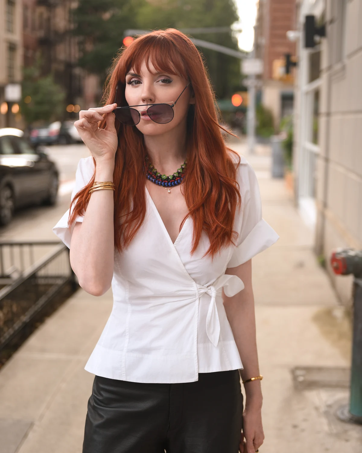 Lindsey in a white blouse, holding her sunglasses on a city sidewalk.